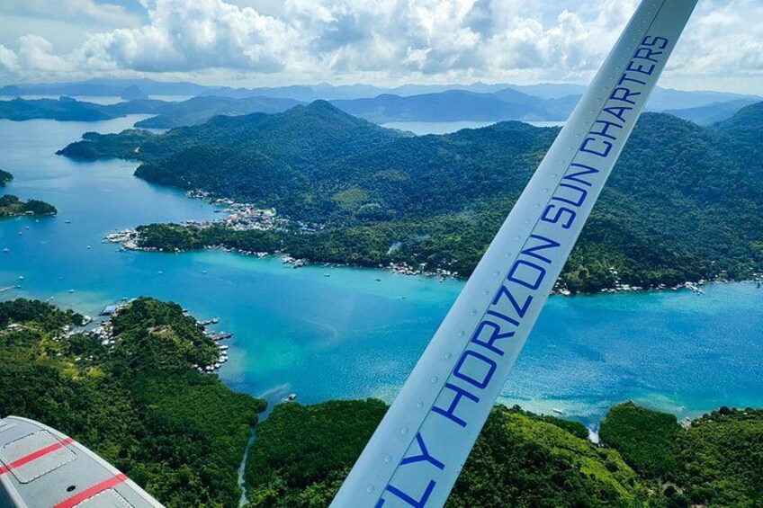 A view of a coastal landscape with clear blue waters, lush green hills, and a visible airplane wing with 'HORIZON SUN CHARTERS' text. 3