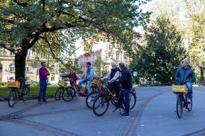People riding bicycles on a paved path in a park with trees and buildings in the background. 5
