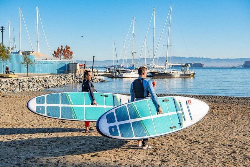 Dos personas cargan tablas de surf en una playa con barcos al fondo. 3