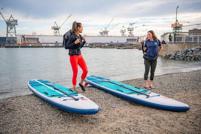 Två personer på en stenstrand med surfbrädor, ett vattendrag och industriella byggnader i bakgrunden. 1