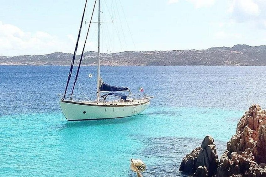 Een zeilboot op een kalme zee met helderblauw water, een rotsachtige kustlijn en heuvels in de verte. 3