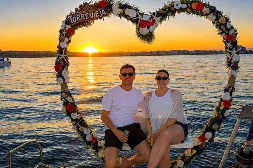 A couple sitting in a boat with a heart-shaped floral arch and a sunset in the background. 3