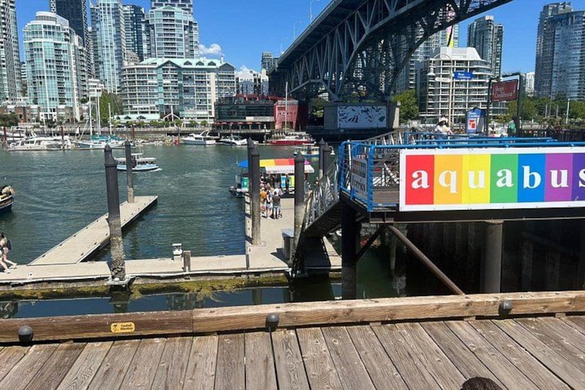 A waterfront with a wooden dock, a colorful sign reading 'aquabus,' and a bridge in the background. 3