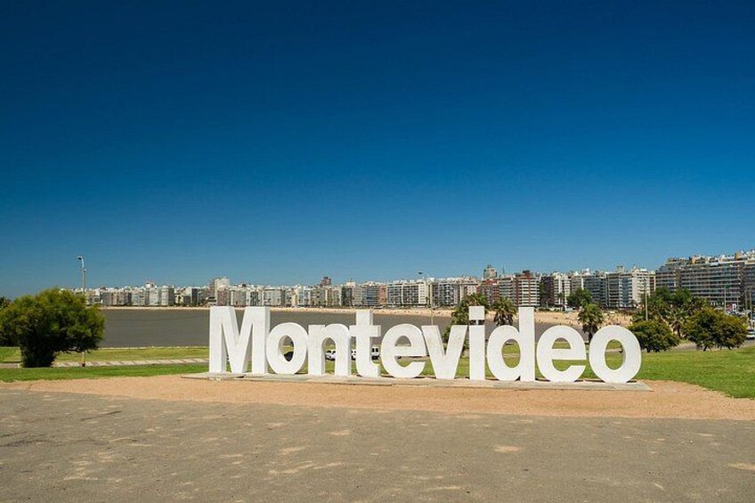 En stor, vit skylt med ordet ”Montevideo” på, placerad i en stadsbild med byggnader och en flod i bakgrunden. 2