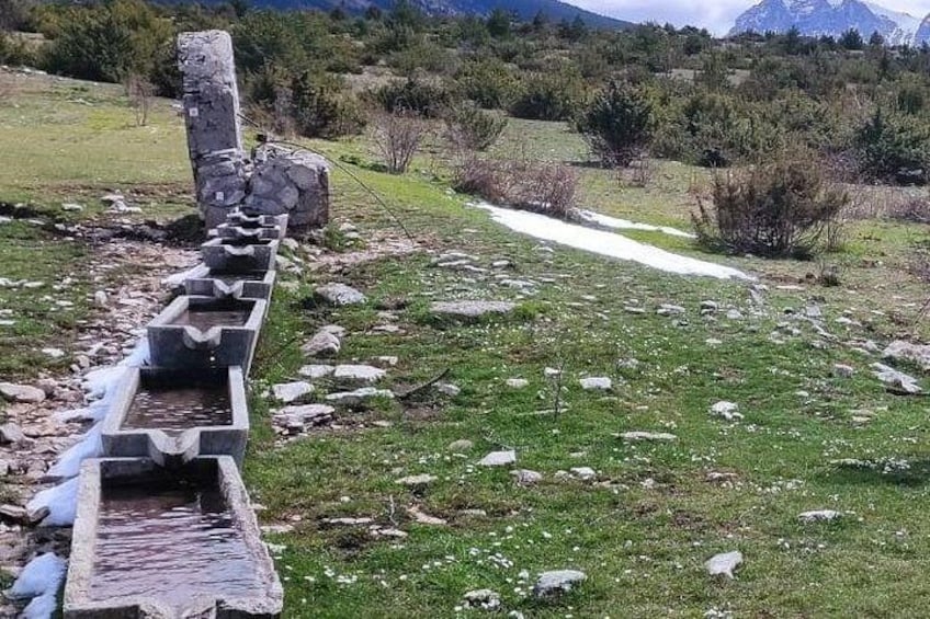 Un canal d’eau en pierre, avec une structure en pierre à son extrémité, entouré d’un champ herbeux et bordé de montagnes en arrière-plan. 1