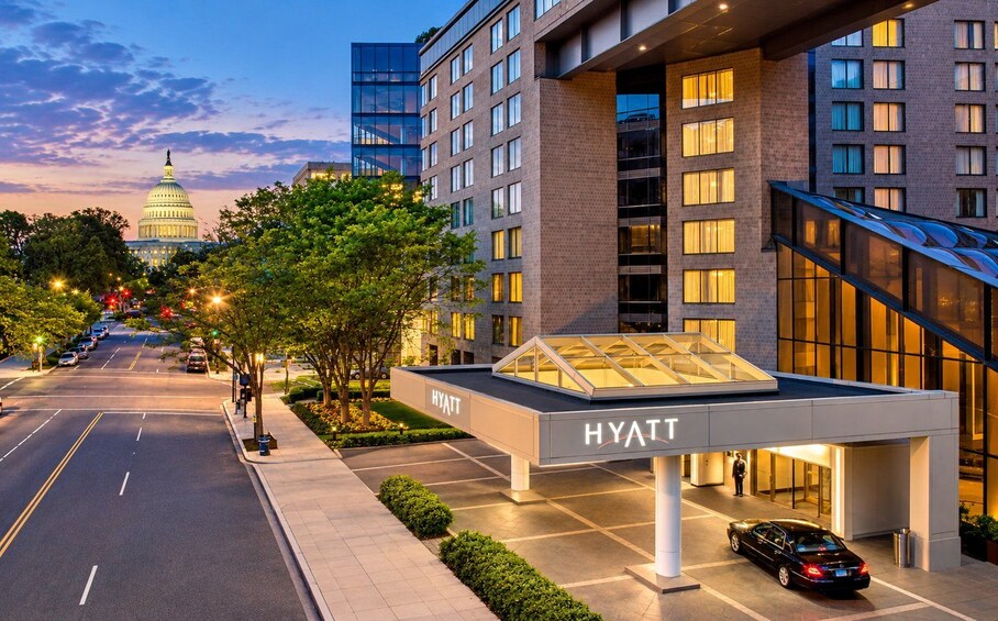 A hotel with a glass canopy entrance, a road with a car, and the Capitol building in the background. 5