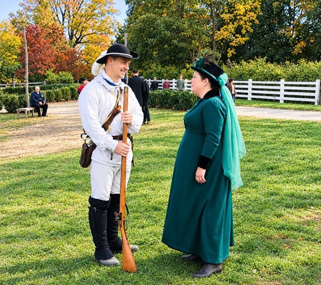 A person in historical attire holding a rifle, standing on grass with trees and a white fence in the background. 3