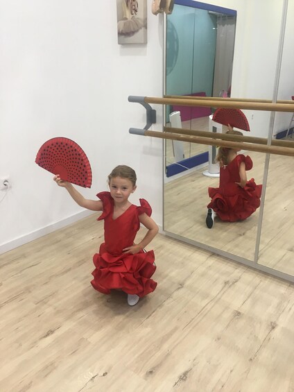 A child in a red dress with a ruffled skirt standing in a room with a large mirror and a ballet barre. 1