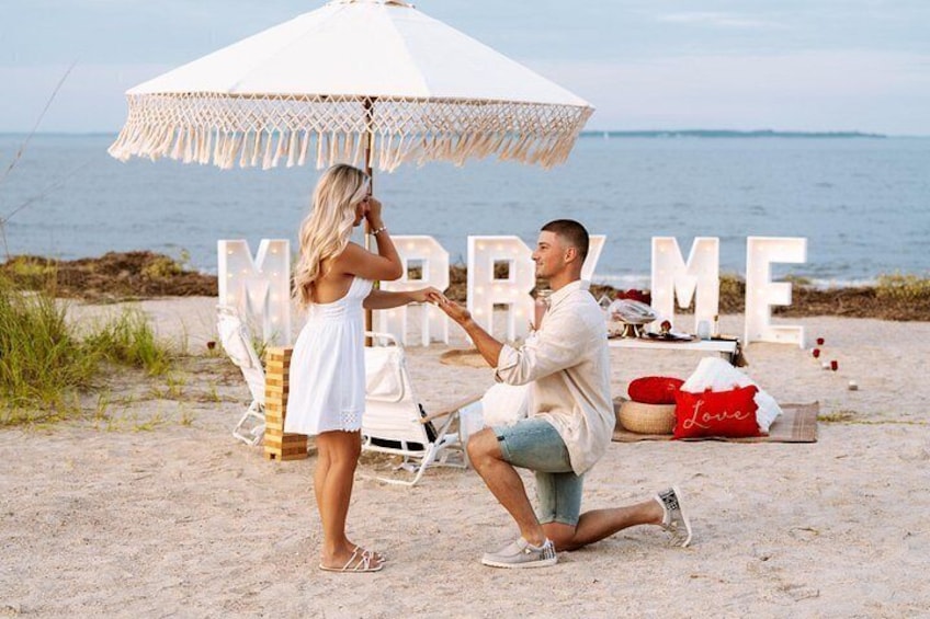 Ein Strandbild, auf dem ein Mann einer Frau unter einem großen Sonnenschirm einen Heiratsantrag macht. 2