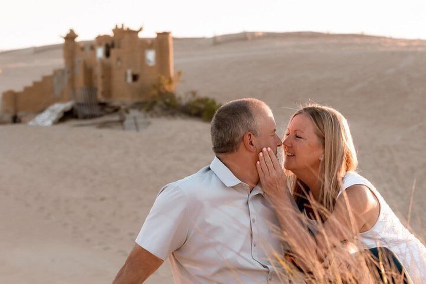 A couple embracing in a desert setting with a historical building in the background. 2