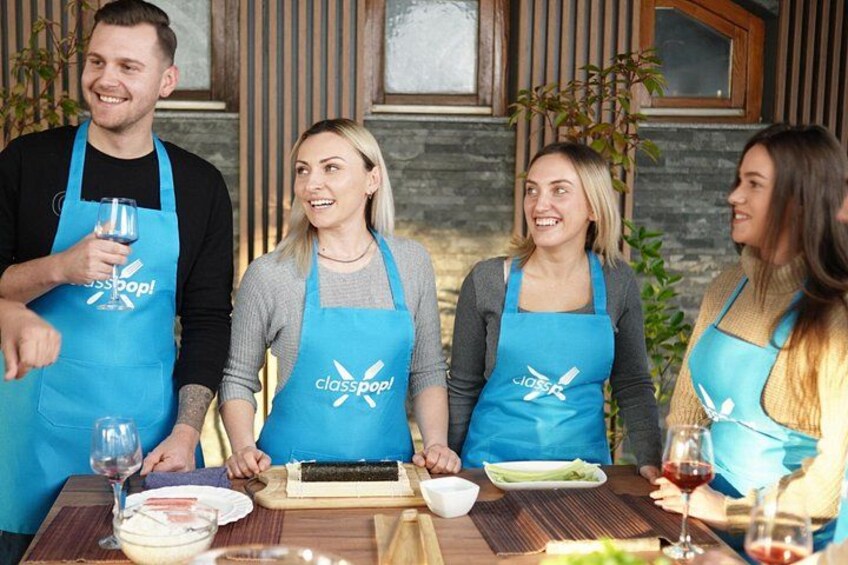 Cuatro personas con delantales azules con el logo 'classpop!' sentadas alrededor de una mesa de madera con comida y vino. 2