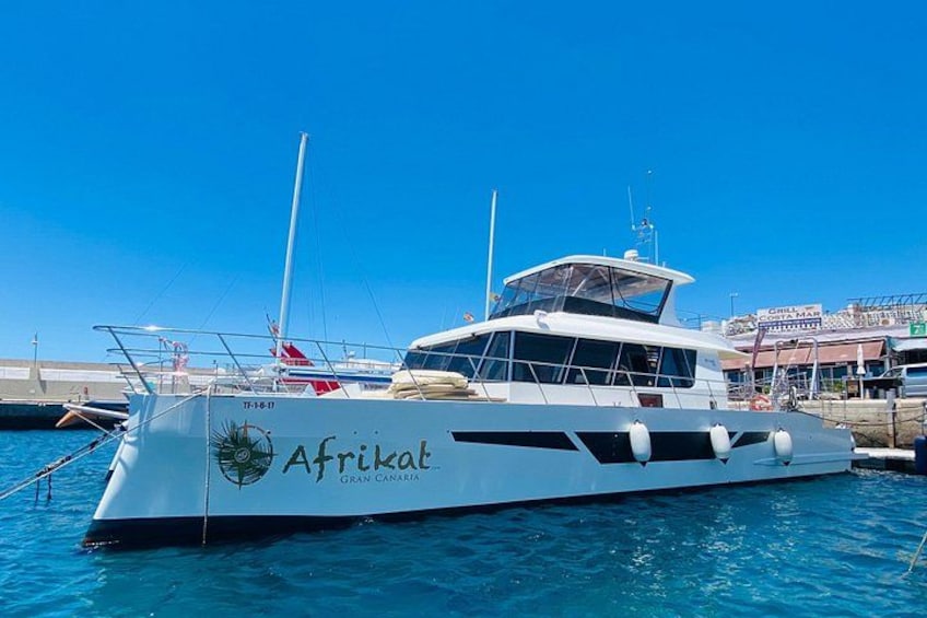 En hvid sejlbåd ved navn Afrikat med et logo, ligger ved et havnemole med krystalklart vand og en klar himmel. 3