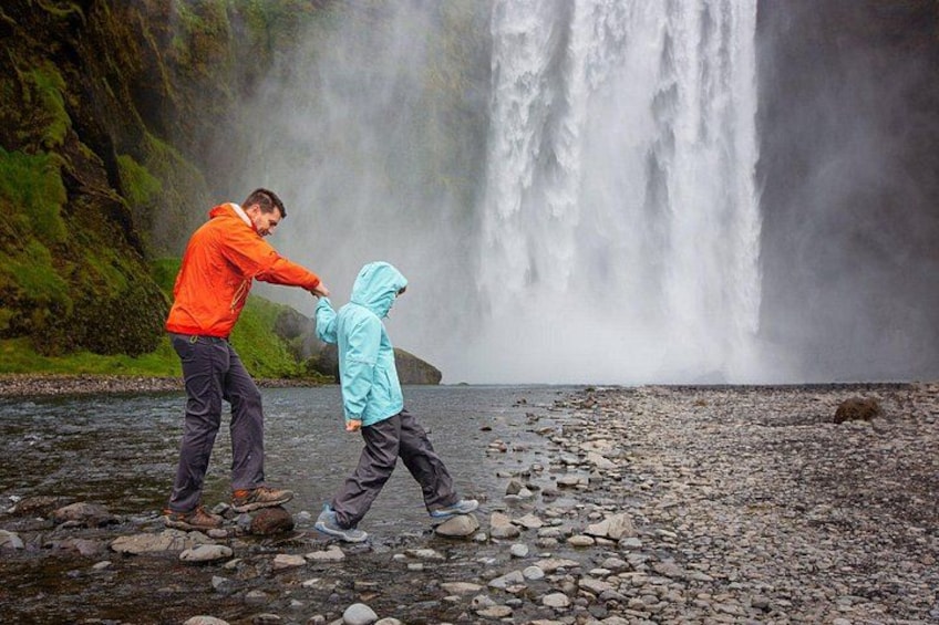 A person in an orange jacket and a child in a blue jacket walking on a rocky shore with a waterfall in the background. 4