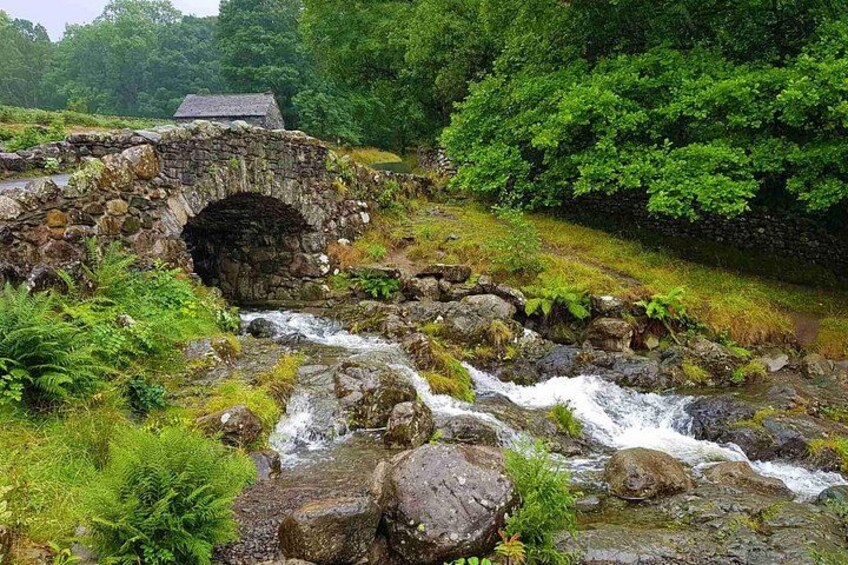 A stone bridge over a flowing stream, surrounded by lush greenery and a stone wall. 5