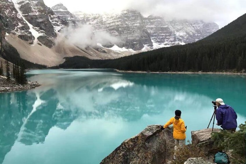 Eine Person in einer gelben Jacke und eine andere in einer blauen Jacke stehen auf einem Felsvorsprung mit Blick auf einen türkisblauen See und Berge im Hintergrund. 2