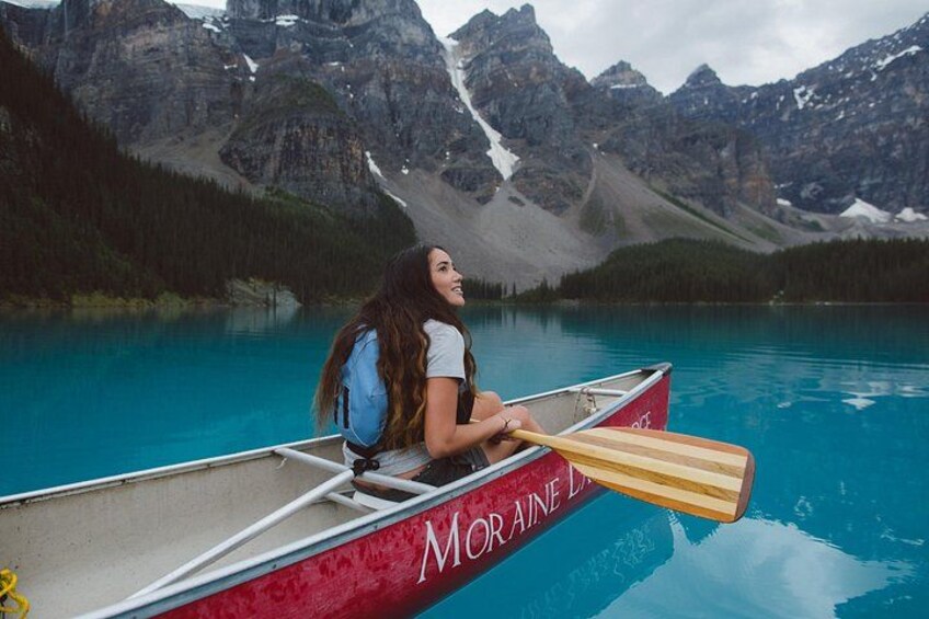 Eine Person in einem roten Kanu mit der Aufschrift 'MORAINE', paddelt auf einem türkisblauen See vor einer Bergkulisse. 1