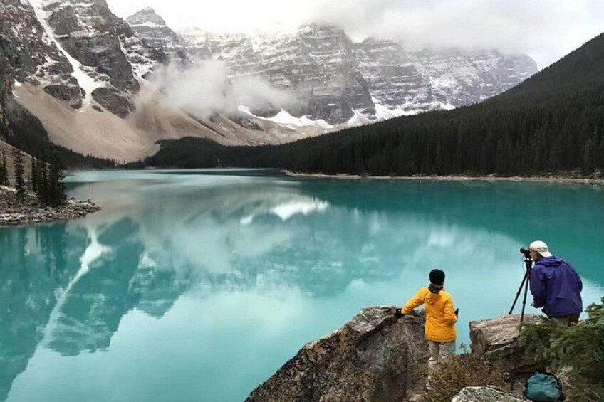 A turquoise lake surrounded by mountains with a person in a yellow jacket and another with a camera on a tripod. 2