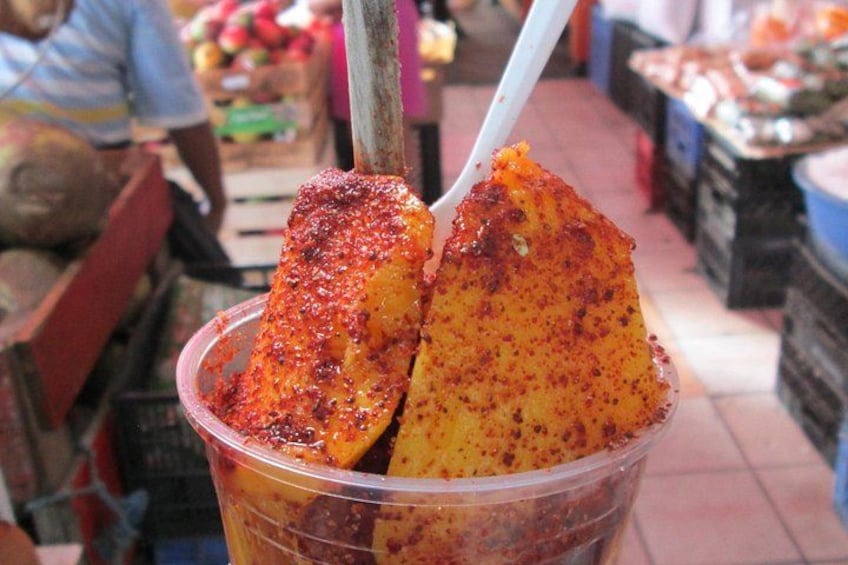 Un bicchiere di plastica con salsa rossa e un frutto giallo, probabilmente mango, in un mercato. 3