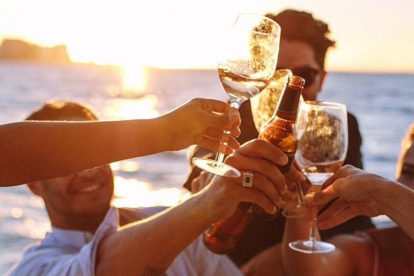 Menschen stoßen mit Champagnergläsern an, eine Flasche steht bereit, während die Sonne über dem Wasser untergeht. 1