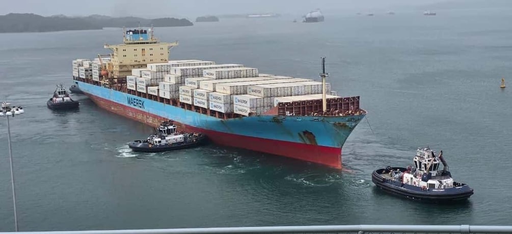 A large cargo ship named 'MAERSK' with multiple containers, being assisted by tugboats in a body of water. 5
