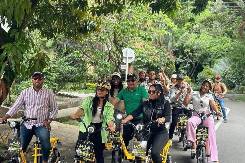 A group of people on bicycles and motorcycles, some wearing helmets, in a green, tree-lined area. 3