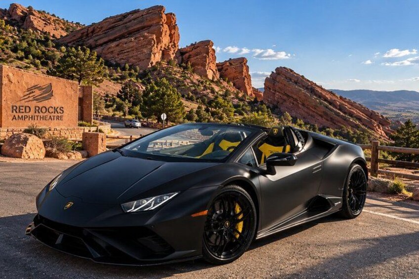 A black sports car parked in front of a rocky landscape with a sign for Red Rocks Amphitheatre. 1