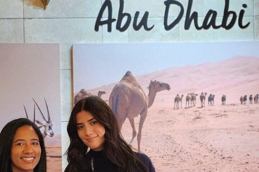 Un camello y un grupo de camellos en un paisaje desértico con un cartel que dice 'Abu Dhabi'. 4