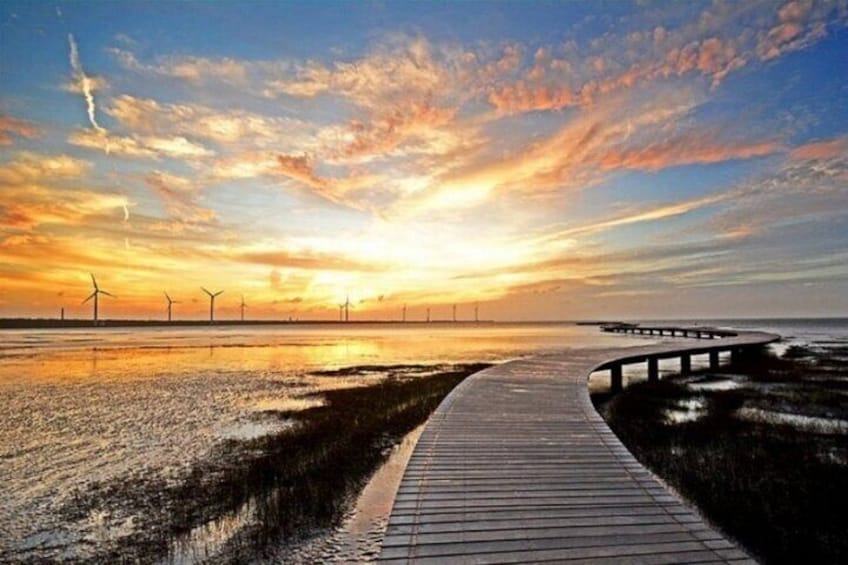 Une promenade en bois qui mène à un point de vue sur un coucher de soleil au-dessus d’une étendue d’eau, avec des éoliennes visibles au loin. 5