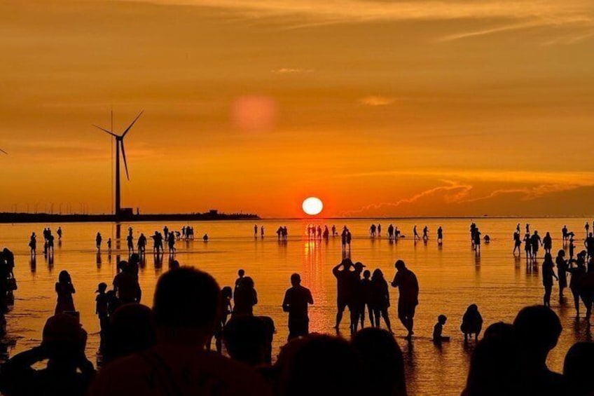 Un atardecer sobre una playa con gente de pie en el agua y aerogeneradores al fondo. 1