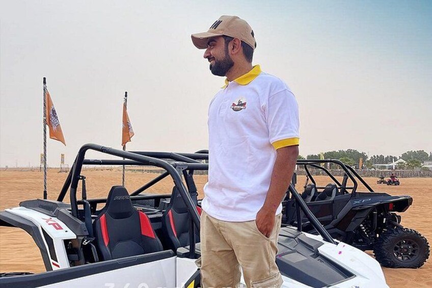 A man standing next to a dune buggy in a desert with flags in the background. 3