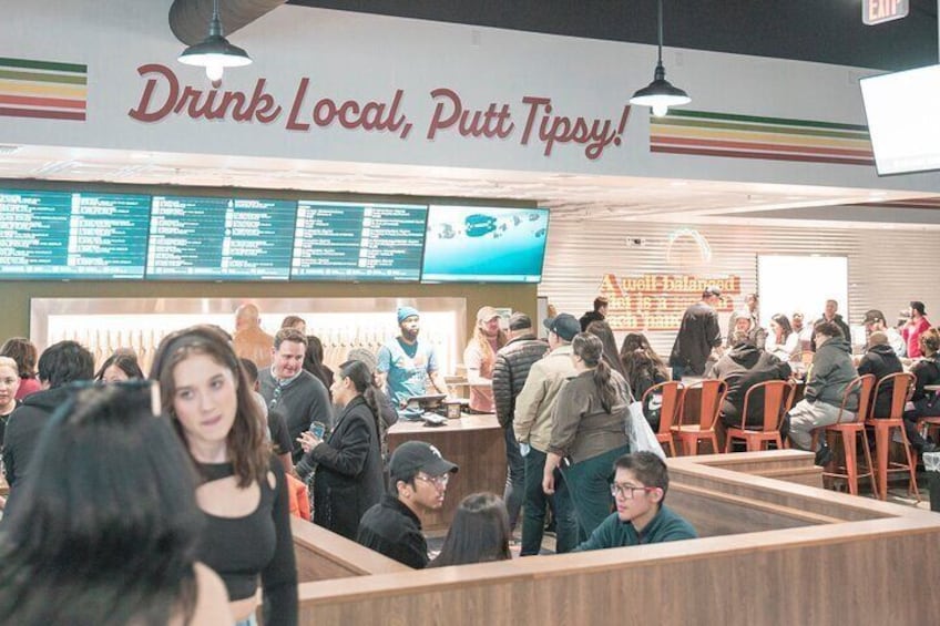 A busy fast-food restaurant with a sign reading "Drink Local, Putt Tipsy!" and a menu board above the counter. 5
