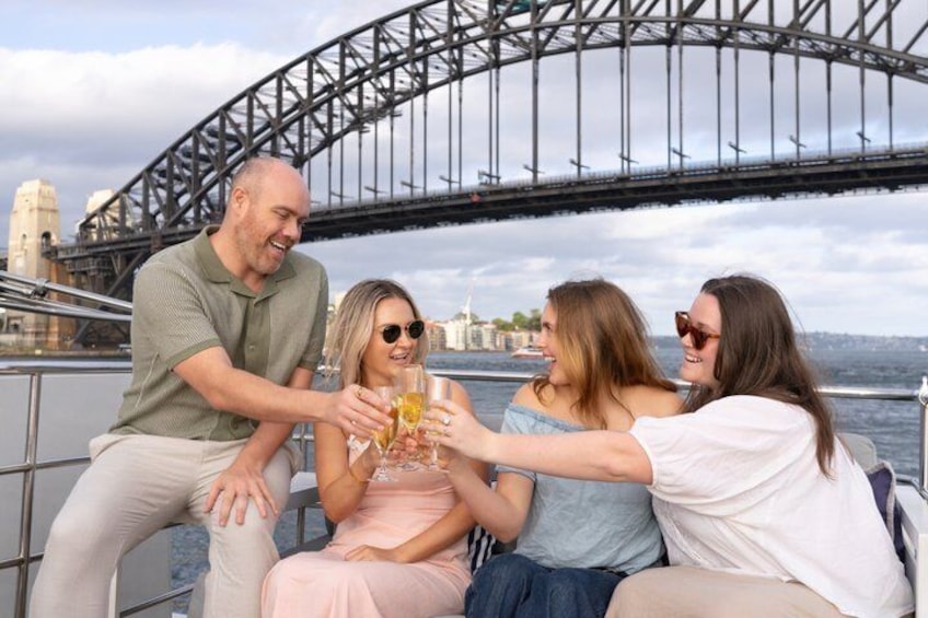 Un grupo de personas brindando con bebidas en un barco, con el Puente de Sydney al fondo. 3
