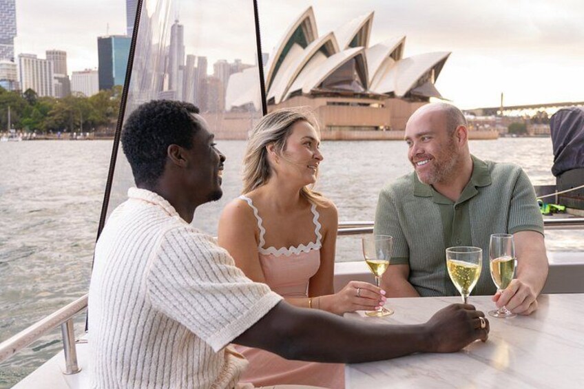 Drie mensen zitten aan een tafel met drankjes, met de Sydney Opera House op de achtergrond. 5