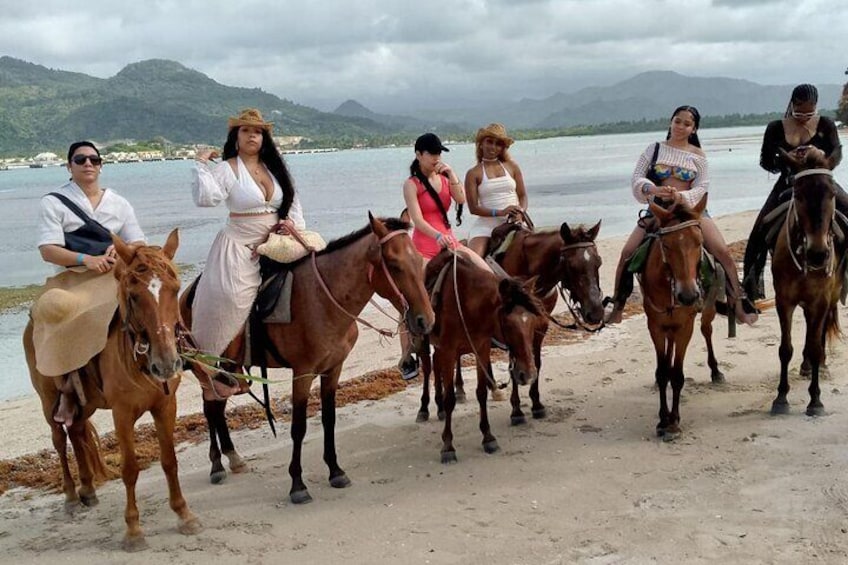 Un gruppo di persone cavalca lungo una spiaggia, con un corpo d'acqua e montagne sullo sfondo. 2