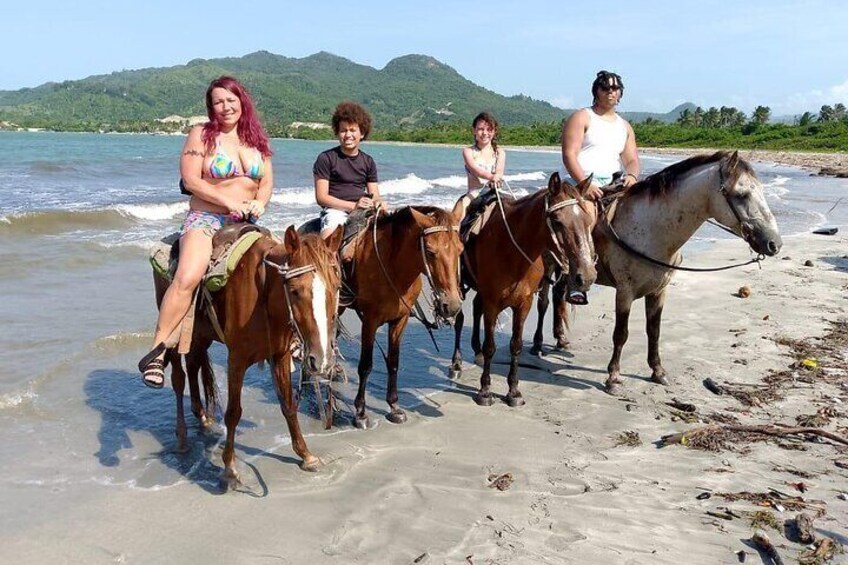 Quattro persone cavalcano a cavallo su una spiaggia, con l'acqua e delle colline sullo sfondo. 4