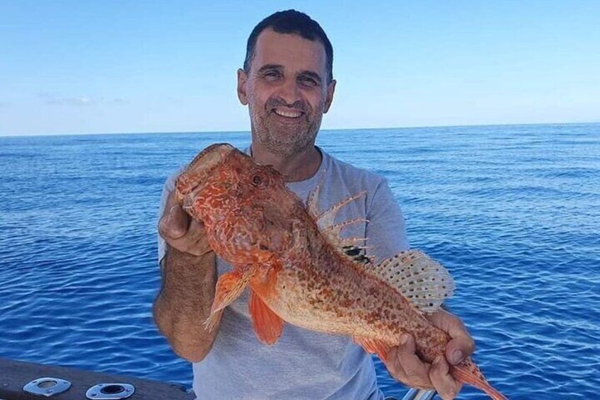 Une personne tenant un gros poisson aux motifs tachetés, debout sur un bateau, avec une étendue d’eau bleue et calme en arrière-plan. 1
