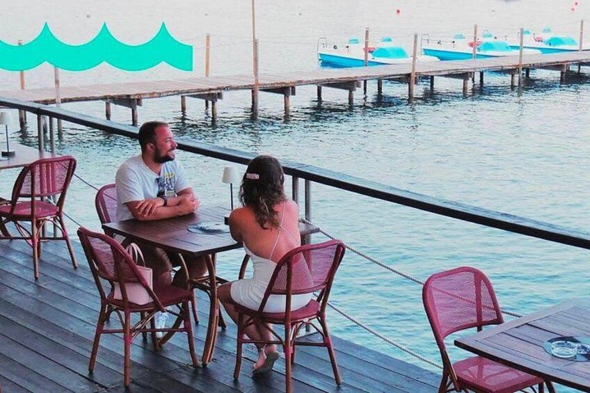 A couple dining at a wooden deck overlooking water with boats in the background. 2