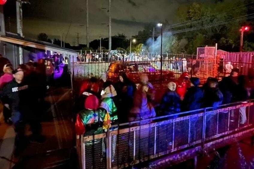 Una multitud de personas en un evento al aire libre, con una cerca y luces de colores. 4
