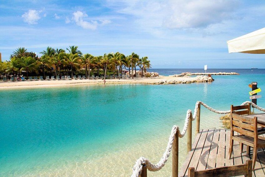 A beach with clear turquoise water, palm trees, and a wooden deck with a rope barrier. 1
