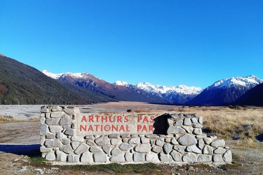 Une pancarte en pierre sur laquelle est inscrit « Parc national d’Arthur’s Pass », avec en arrière-plan des montagnes et un ciel bleu clair. 3
