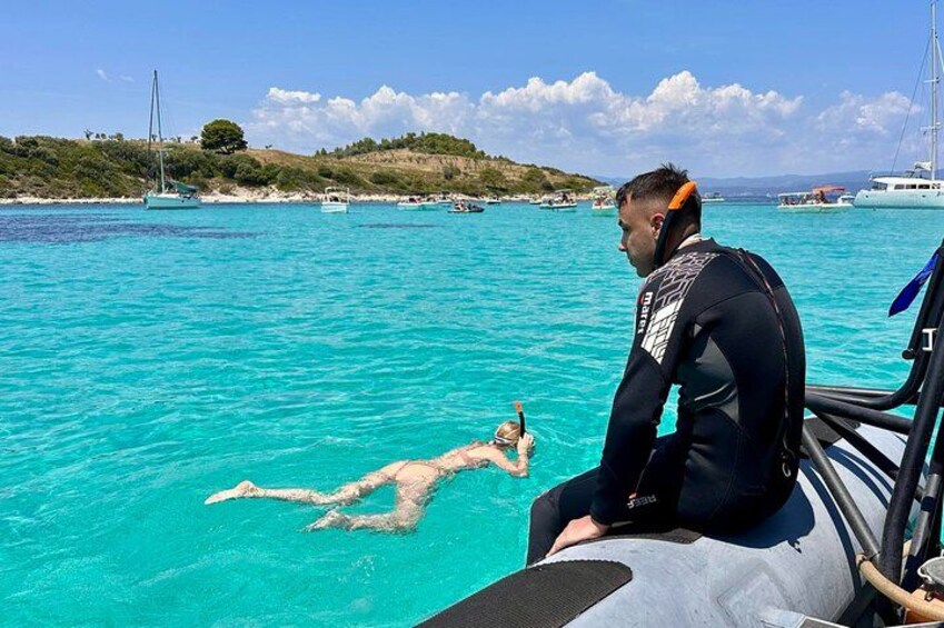 A person snorkeling in clear blue water, a boat with a person on board, and a distant shoreline with boats and hills. 3