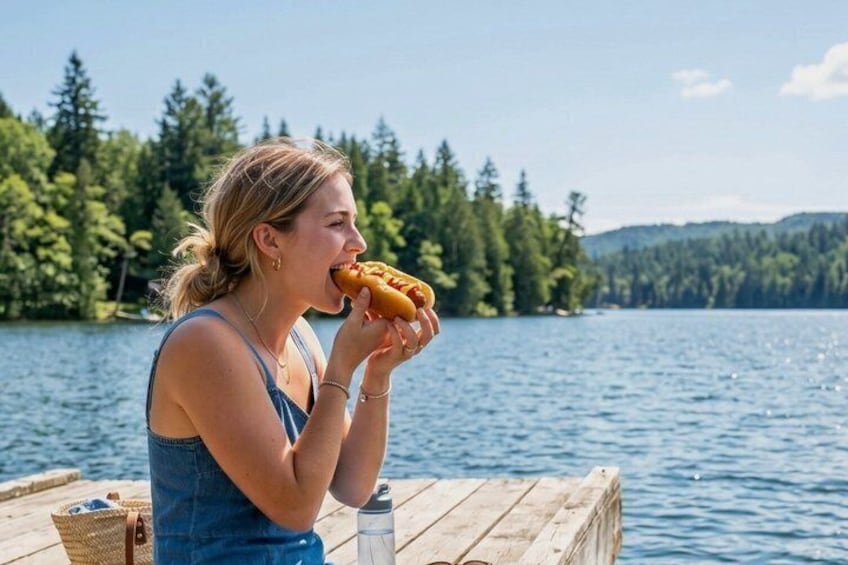 Una persona comiendo un perro caliente en un muelle de madera junto a un lago, con árboles al fondo. 5