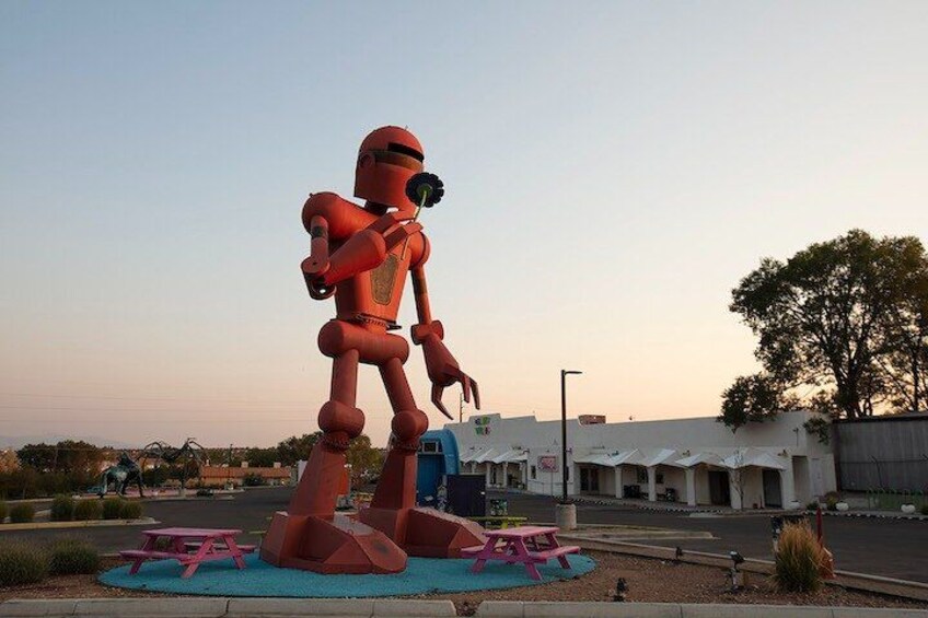 A large red robot sculpture in an outdoor setting with a parking lot and buildings in the background. 2