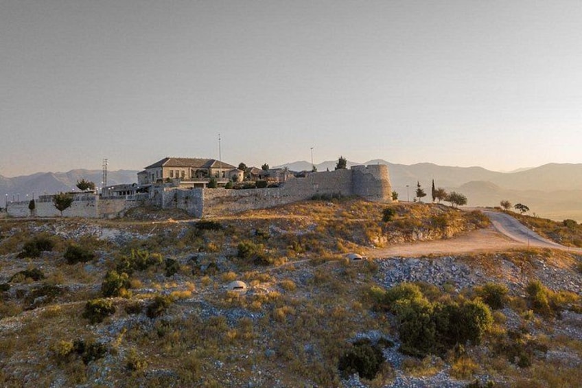 A historic building on a hill with a stone wall, a dirt path, and sparse vegetation. 1