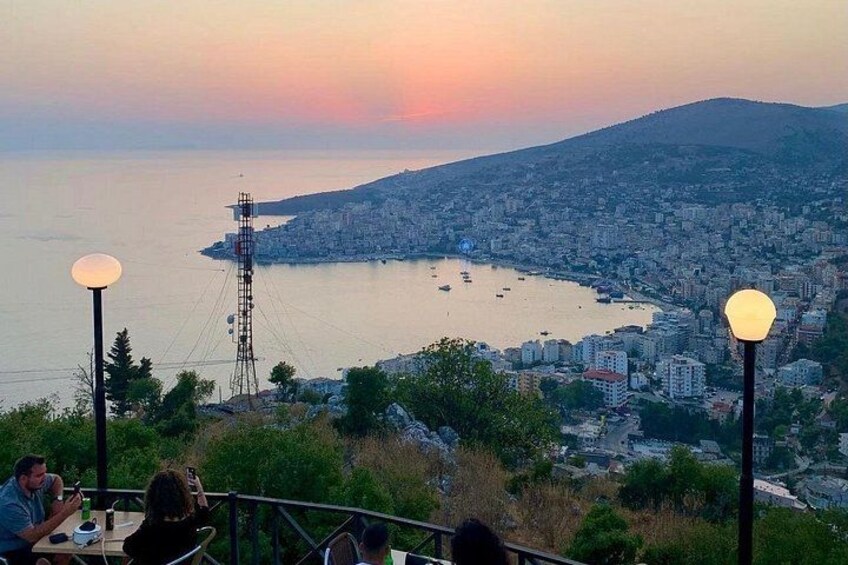 A coastal cityscape at sunset with a prominent communication tower, a body of water with boats, and a densely built urban area. 4
