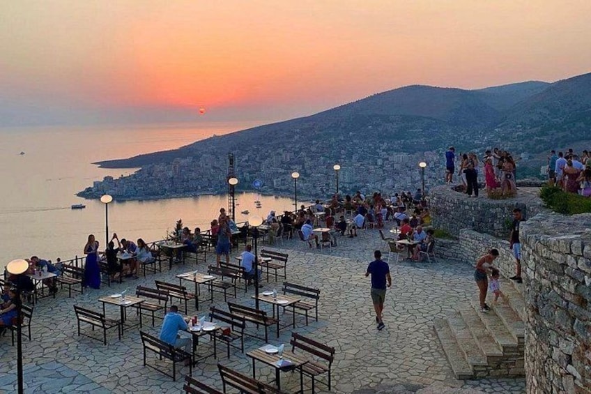 A sunset view with people dining at outdoor tables, a stone wall, and a cityscape in the background. 5