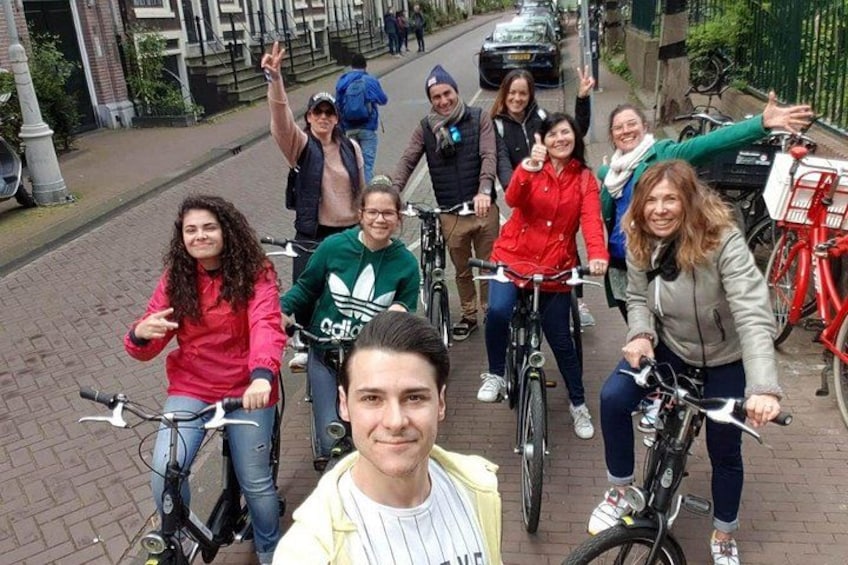 A group of people on bicycles on a paved street with buildings and parked bicycles in the background. 4