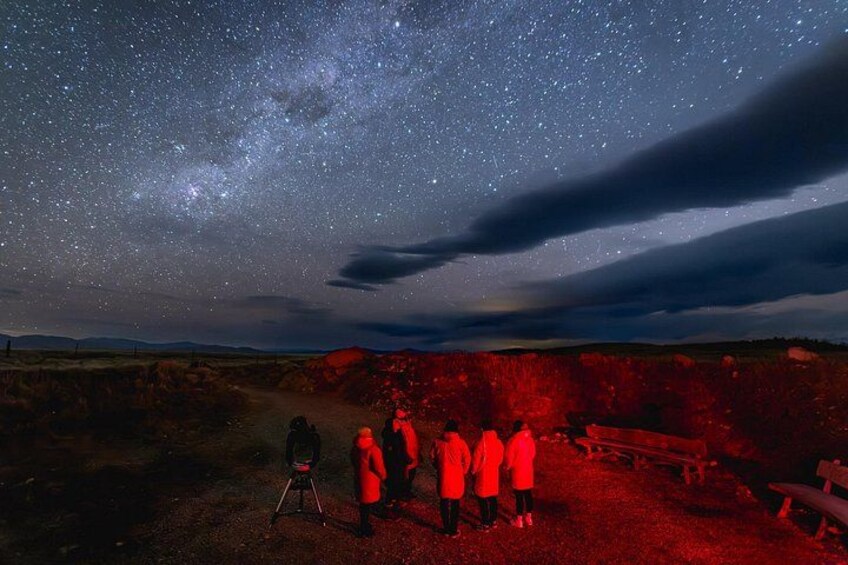 A starry night sky with the Milky Way visible, a group of people observing the sky, and a red-lit landscape. 1