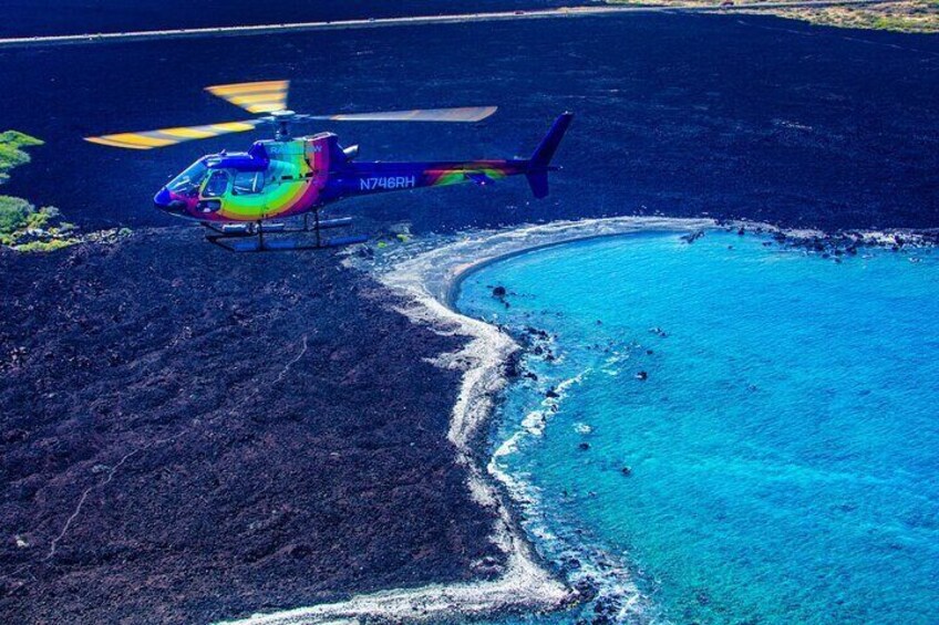 A helicopter with registration N746RH hovering over a coastline with clear blue waters and dark volcanic terrain. 2