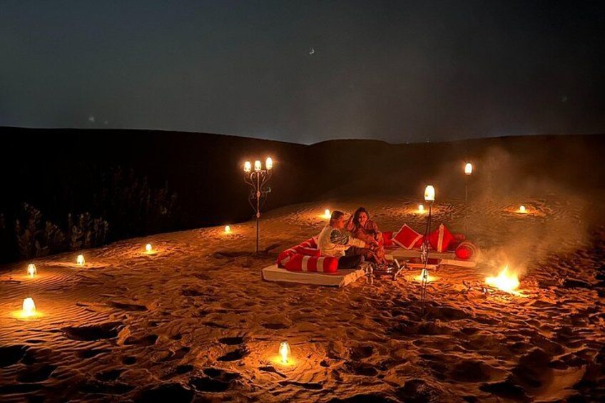 A desert campsite at night with people sitting around a fire, surrounded by lit candles and a candelabra. 2